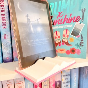 May include: A Kindle e-reader with a pink case rests on a white and pink book-shaped stand. The screen displays text: "Your next great read is waiting." Bookshelves with colorful book spines are in the background.