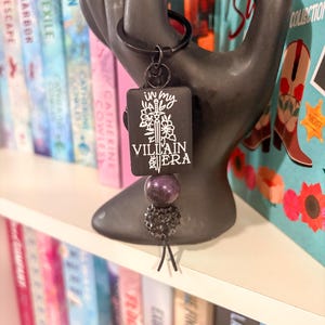 Puede incluir: Llavero negro con un colgante rectangular con el texto blanco "IN MY VILLAIN ERA" y un diseño de espada y flores. El llavero tiene un anillo negro y está adornado con cuentas moradas y negras. El fondo incluye libros coloridos.