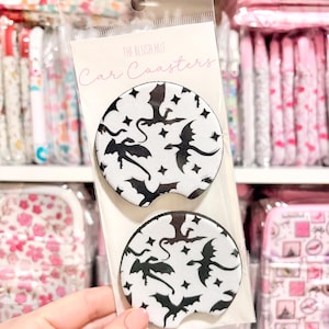 May include: Two car coasters with a white background and a black dragon and star pattern. The coasters are packaged in a clear plastic sleeve with the text "THE BLUSH HUT Car Coasters". The coasters are round and designed to fit in a car's cup holder.