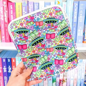 Op de afbeelding: Een kleurrijke tablethoes met een herhalend patroon van UFO's, boeken, harten, sterren en cadeau dozen. De hoes heeft een witte rits en rand. De achtergrond is wit.