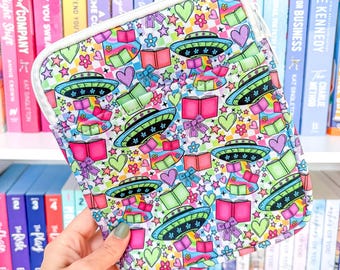 Puede incluir: Una funda de tableta colorida con un patrón repetido de ovnis, libros, corazones, estrellas y cajas de regalo. La funda tiene una cremallera y un ribete blancos. El fondo es blanco.