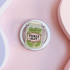 May include: A clear, circular phone grip featuring a jar of pickles design. The jar has a beige lid and a label that reads "PICKLE SLUT" in pink and beige. The pickles are green.