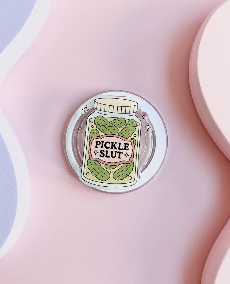 May include: A clear, circular phone grip featuring a jar of pickles design. The jar has a beige lid and a label that reads "PICKLE SLUT" in pink and beige. The pickles are green.