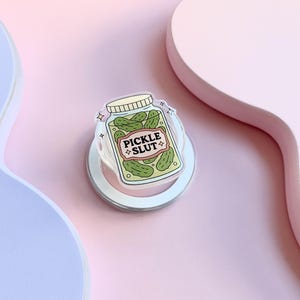 May include: A clear phone grip featuring a jar of pickles with the text "PICKLE SLUT". The jar is filled with green pickles and has a white lid. The grip is set against a pink backdrop.