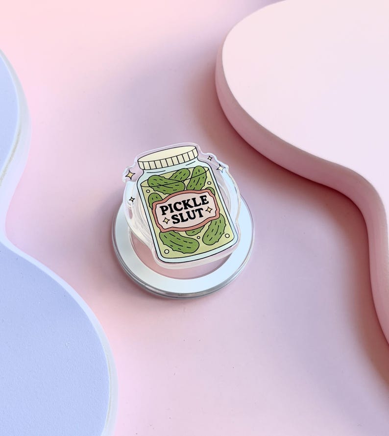 May include: A clear phone grip featuring a jar of pickles with the text "PICKLE SLUT". The jar is filled with green pickles and has a white lid. The grip is set against a pink backdrop.