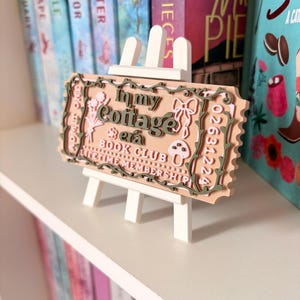 May include: A peach-coloured book club sign with the text "In my Cottage Book Club" and "Lifetime Membership", displayed on a small white easel. The sign has green and pink decorative elements, including flowers and bows. The sign is placed near a bookshelf.