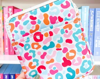 Puede incluir: Una funda blanca con cremallera y un colorido diseño de estampado de leopardo en tonos rosa, naranja y azul. La funda tiene un borde blanco y un bolsillo frontal. La funda está diseñada para guardar libros u otros artículos.