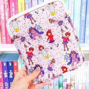 Può includere: Una custodia rosa per laptop con cerniera bianca e un motivo di ragazze dei cartoni animati in vari abiti e pose, con punti di coriandoli colorati. La custodia è tenuta davanti a una libreria.