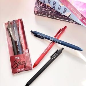 May include: A pen set with the text "The Spicy Pen Set" on the packaging. The set includes black, gray, and red pens with phrases like "Filthy" and "Plot Twist: Its Smut". Several pens are scattered around the set. Books are in the background.