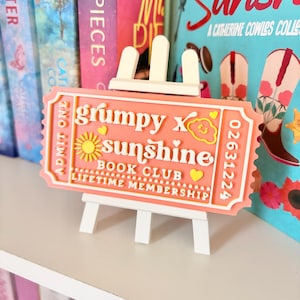 May include: A peach-colored ticket-shaped sign with the words "grumpy x sunshine BOOK CLUB LIFETIME MEMBERSHIP" in white and yellow, displayed on a small white easel. The sign has a sun and cloud design. Books are in the background.