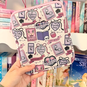 May include: A rectangular tablet sleeve with a white background featuring a repeating pattern of purple and pink illustrations including handcuffs, books, skulls, phones with text like Villain Lover, Kidnapped, and Morally Grey, finished with pink piping.