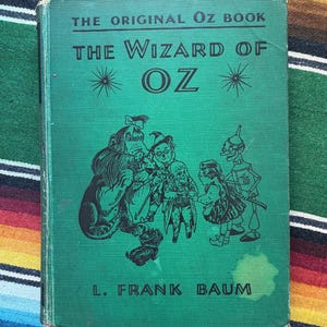May include: A green hardcover book titled The Original Oz Book The Wizard of Oz by L. Frank Baum. The cover features black line art illustrations of the Cowardly Lion, Scarecrow, Dorothy, and the Tin Woodman, with starburst accents above the title.