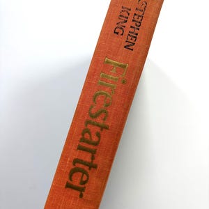 May include: A close-up of the spine of a hardcover book titled "Firestarter" by Stephen King. The book's spine is a burnt orange color with gold lettering. The book is angled against a white background.
