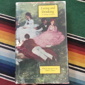 May include: A book titled "Eating and Drinking: An Anthology for Epicures" with an illustration of a group of people having a picnic in a garden. The book cover features a yellow label with the text "With an Introduction by Andre L. Simon".