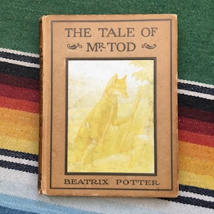 May include: A vintage copy of "The Tale of Mr. Tod" by Beatrix Potter. The book has a tan cover with the title and author's name in brown lettering. The cover features a yellow illustration of a fox. The book is resting on a striped textile.
