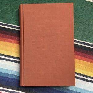 May include: A closed, hardback book with a textured, reddish-brown cover. The book is positioned vertically, with the spine on the left. The background is a colorful striped textile with green, blue, yellow, and red hues.