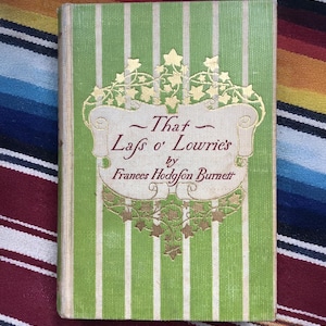 May include: A vintage book titled "That Lass o' Lowrie's" by Frances Hodgson Burnett. The cover features a green and white striped pattern with gold decorative elements and text. The book's cover is slightly worn.