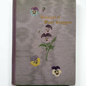 May include: A hardcover book with a textured grey cover featuring a central illustration of purple and yellow pansy flowers. The title Rubaiyat of Omar Khayyam is printed in gold lettering on the front cover. The book has a dark purple spine.
