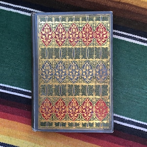 May include: A vintage book with a decorative cover. The cover features a repeating pattern of red and blue floral designs with gold accents on a dark green background. The book's spine is a dark gray color.