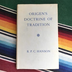 May include: A vintage book titled "Origen's Doctrine of Tradition" by R. P. C. Hanson. The book has a light blue cover with blue text and a small floral design. The book is resting on a colorful striped textile.