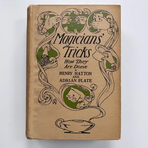 May include: A tan hardcover book titled Magicians' Tricks How They Are Done by Henry Hatton and Adrian Plate. The cover features black line art illustrations of hands performing magic tricks, green circular accents, and a lamp at the bottom.