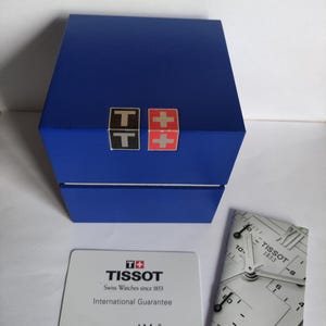 Może przedstawiać: Niebieskie pudełko na zegarek z logo Tissot i kartą gwarancyjną. Pudełko ma dwa małe logo na górze. Karta gwarancyjna zawiera logo Tissot i tekst "Swiss Watches since 1853". Obecny jest również obraz tarczy zegarka.