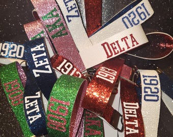 May include: A collection of glittery wrist keychains in various colors, including red, green, pink, white, and blue. Each keychain features text such as "ZETA", "DELTA", and numbers like "1913" and "1920".