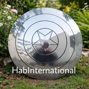 May include: A large, shiny, silver shield with a central star design and concentric circles. The shield has a reflective surface and is displayed outdoors on grass. The text "HablInternational" is visible at the bottom.