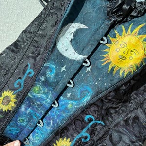 Peut inclure: Un corset noir avec un motif damass&eacute; textur&eacute; et un panneau central peint &agrave; la main. Le design pr&eacute;sente un croissant de lune blanc, un soleil jaune vif avec un visage, des motifs tourbillonnants bleus et des tournesols sur fond bleu fonc&eacute;.