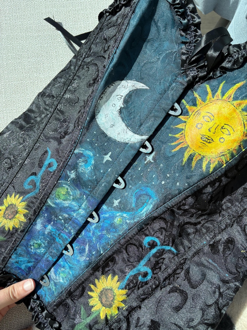 Peut inclure: Un corset noir avec un motif damass&eacute; textur&eacute; et un panneau central peint &agrave; la main. Le design pr&eacute;sente un croissant de lune blanc, un soleil jaune vif avec un visage, des motifs tourbillonnants bleus et des tournesols sur fond bleu fonc&eacute;.