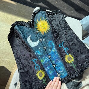 Peut inclure: Un corset noir avec un motif damass&eacute; et un panneau central peint repr&eacute;sentant un soleil jaune, un croissant de lune blanc, des nuages bleus tourbillonnants et deux tournesols jaunes. Le v&ecirc;tement pr&eacute;sente un bord volant&eacute; en haut et en bas.