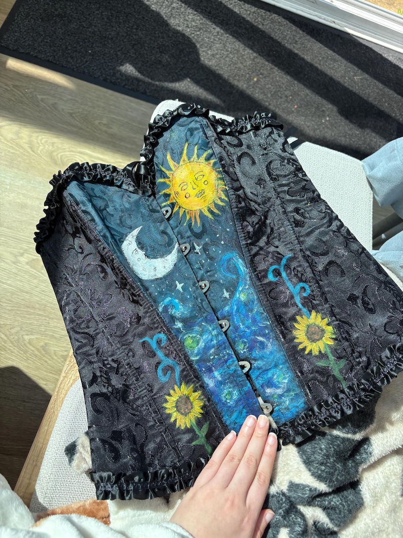 Peut inclure: Un corset noir avec un motif damass&eacute; et un panneau central peint repr&eacute;sentant un soleil jaune, un croissant de lune blanc, des nuages bleus tourbillonnants et deux tournesols jaunes. Le v&ecirc;tement pr&eacute;sente un bord volant&eacute; en haut et en bas.