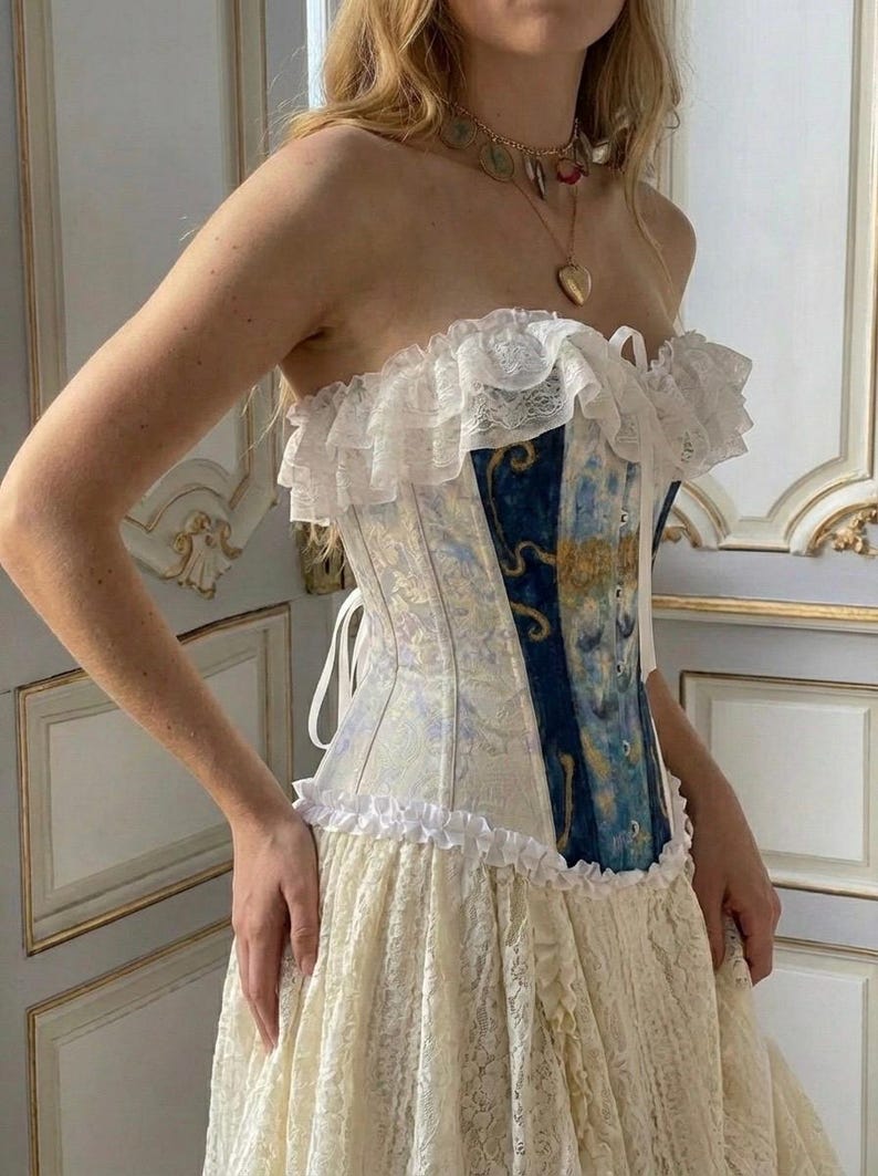 May include: A strapless corset top featuring a mix of cream-colored fabric and a central panel with a blue and gold swirling pattern. The garment is accented with white lace ruffles along the top and bottom edges and includes a white lace skirt.