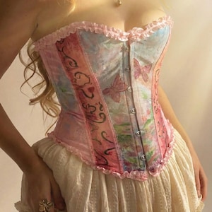 May include: A strapless corset top featuring a pastel colour palette of pink and blue with a butterfly print. The garment has a front button closure, ruffled trim along the top and bottom edges, and vertical paneling with decorative dark line art.