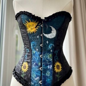 Peut inclure: Un corset bustier orn&eacute; d'un imprim&eacute; c&eacute;leste bleu fonc&eacute; et noir avec un soleil jaune, un croissant de lune blanc et de petites &eacute;toiles. Le v&ecirc;tement pr&eacute;sente deux tournesols jaunes en bas, des bordures &agrave; volants noirs et des fermoirs argent&eacute;s.