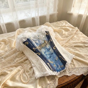 May include: A corset featuring a central panel with a blue and gold celestial print depicting moons and stars, flanked by white side panels with a subtle tonal pattern. The garment is finished with white ruffled lace trim along the top and bottom edges.