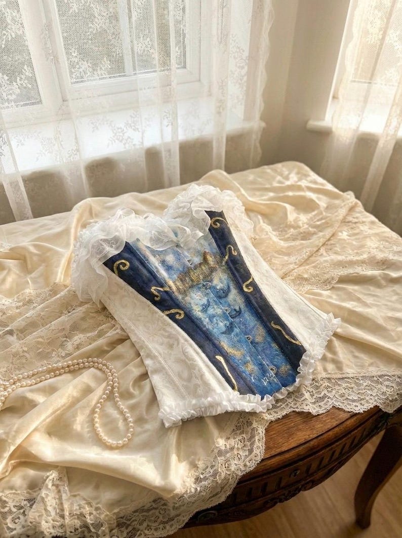 May include: A corset featuring a central panel with a blue and gold celestial print depicting moons and stars, flanked by white side panels with a subtle tonal pattern. The garment is finished with white ruffled lace trim along the top and bottom edges.