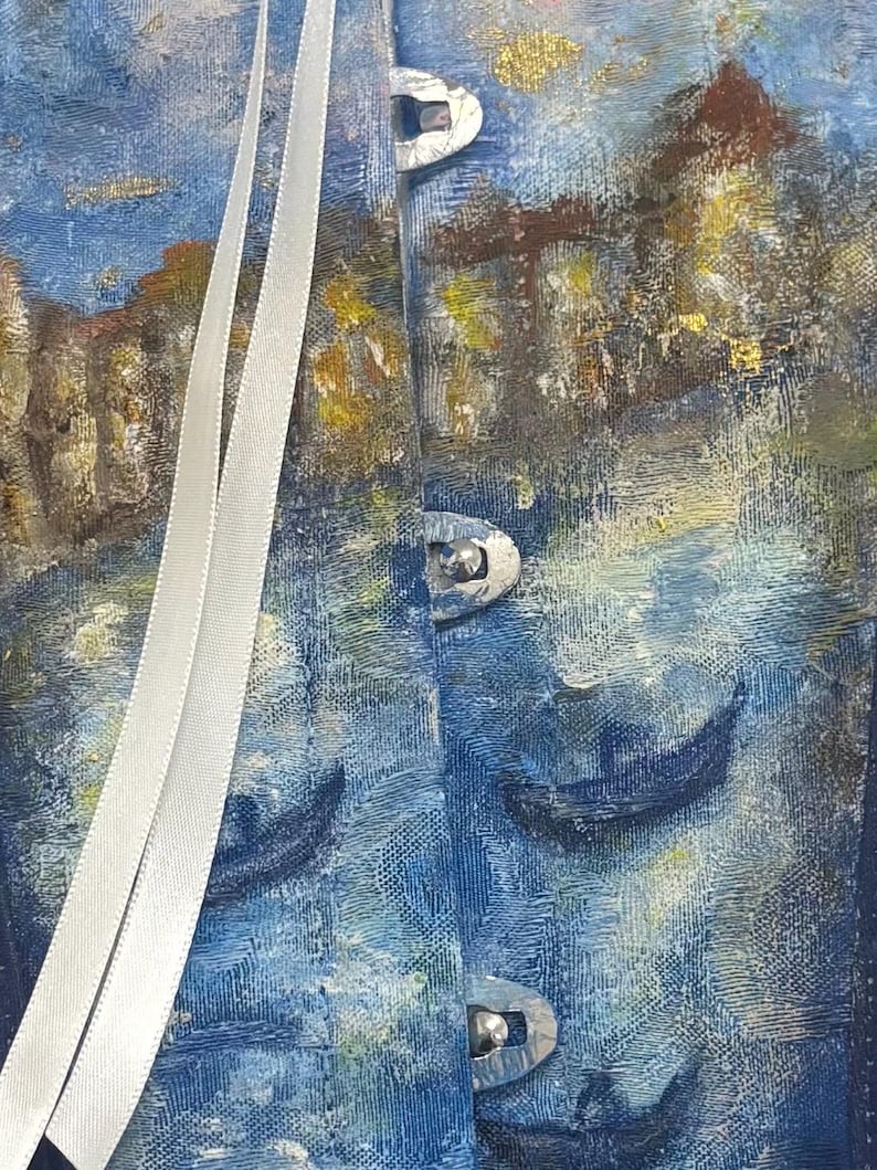 May include: A corset featuring a textured, painted scene of blue water with dark boat silhouettes and golden architectural elements. The garment includes three silver-toned metal front busk closures and two long, smooth white ribbon laces hanging vertically.