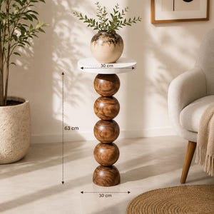 Op de afbeelding: Een ronde witte tafel met een verticale kolom van vijf donkerbruine houten bollen als onderstel. De tafel meet 30 bij 30 bij 63 cm. Op het blad staat een keramieken vaas met een beige en donkerbruin kleurverloop, gevuld met groen blad.