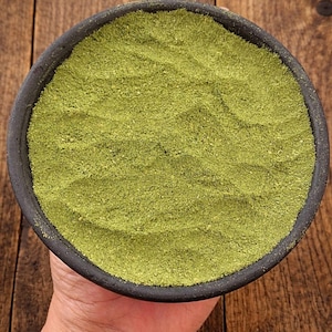 May include: A black bowl filled with finely ground, light green powder, viewed from above. The bowl is held in a hand, set against a wooden background. The powder is likely a culinary ingredient, such as a spice or herb.