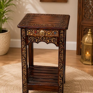 Könnte beinhalten: Ein dunkelbrauner Beistelltisch aus Holz mit aufwendigen Blumenverzierungen an den Beinen und der Schubladenfront. Die Schublade hat einen runden Messinggriff und kleine, farbige Einlegearbeiten. Er verfügt über eine flache Platte und ein Regal.