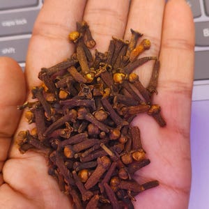May include: A handful of dried cloves, a spice used in cooking and baking. The cloves are a dark brown color with small, round heads and slender stems. The image is taken from a close-up perspective, showing the texture and detail of the cloves.