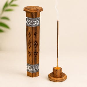 May include: A wooden incense holder and stand set. The tall, rectangular holder has silver floral accents and diamond-shaped cutouts. A lit incense stick emits a wisp of smoke, placed in a matching wooden stand. The set is made of brown wood.