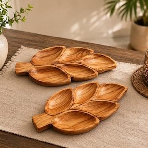 May include: Two wooden serving trays shaped like leaves with distinct grain patterns. Each tray features a central stem and four individual concave sections for holding items. The wood has a warm, light brown tone with natural dark brown streaks.