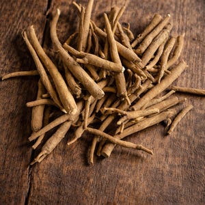 May include: A pile of dried, light brown ashwagandha roots on a dark wooden surface. The roots are cylindrical and vary in size, with some tapering to a point. The wood grain is visible, adding texture to the image.