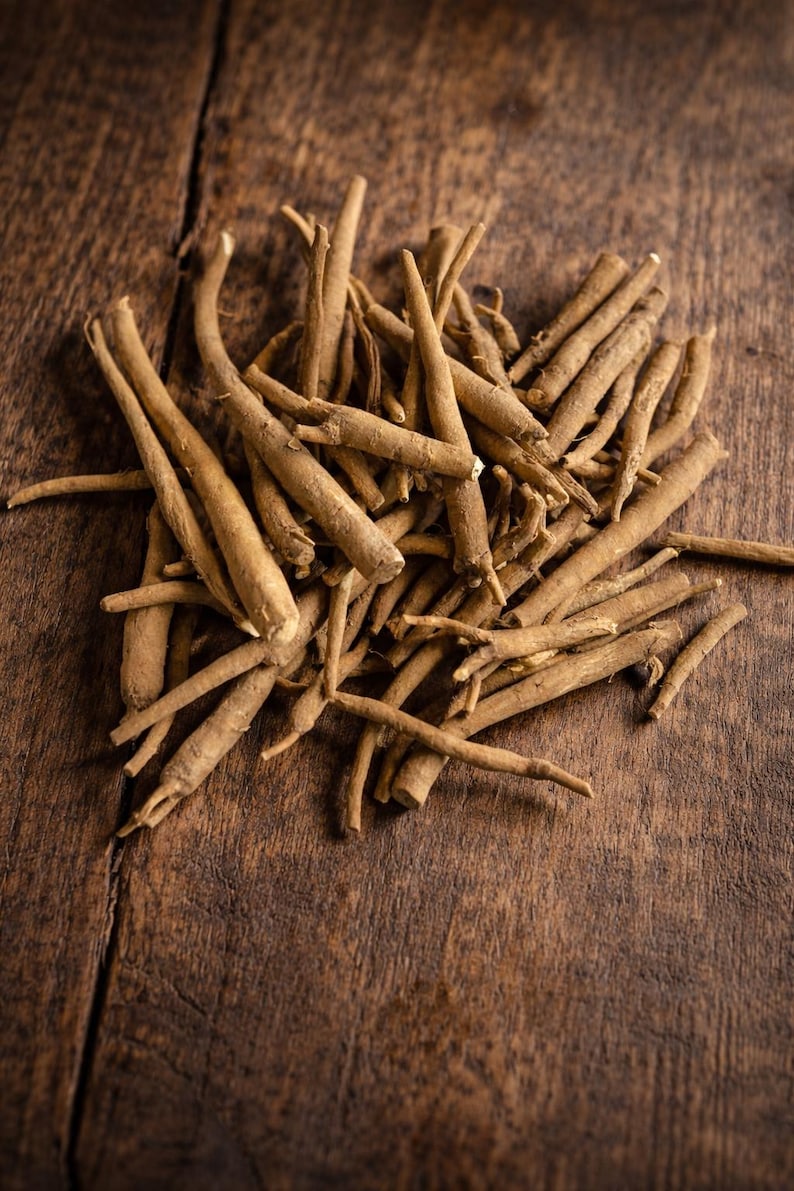 May include: A pile of dried, light brown ashwagandha roots on a dark wooden surface. The roots are cylindrical and vary in size, with some tapering to a point. The wood grain is visible, adding texture to the image.