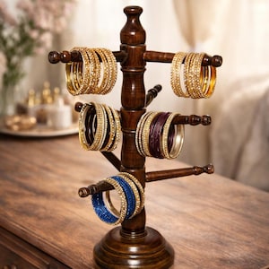 May include: A dark brown wooden bangle stand displaying various gold, purple, and blue bangles. The stand has multiple arms to hold the bangles, and is placed on a wooden surface. The bangles are adorned with decorative elements.
