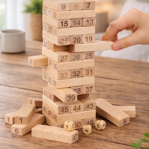 May include: A wooden block stacking game with numbered blocks and dice. The blocks are arranged in a tower, with some blocks removed. A hand is reaching for a block. The game is on a wooden table.