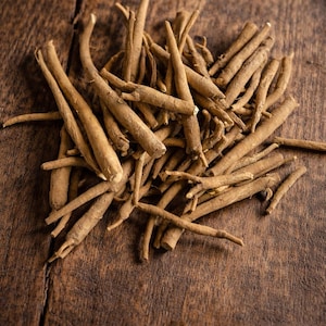 May include: A pile of dried, light brown ashwagandha roots on a dark wooden surface. The roots are cylindrical and vary in size, with some tapering to a point. The wood grain is visible, adding texture to the image.