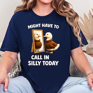 以下が含まれることがあります： 鏡を見ている漫画のアヒルのネイビーブルーのTシャツ。アヒルとその反射は茶色のセーターを着ています。シャツのテキストは「MIGHT HAVE TO CALL IN SILLY TODAY」と書かれています。